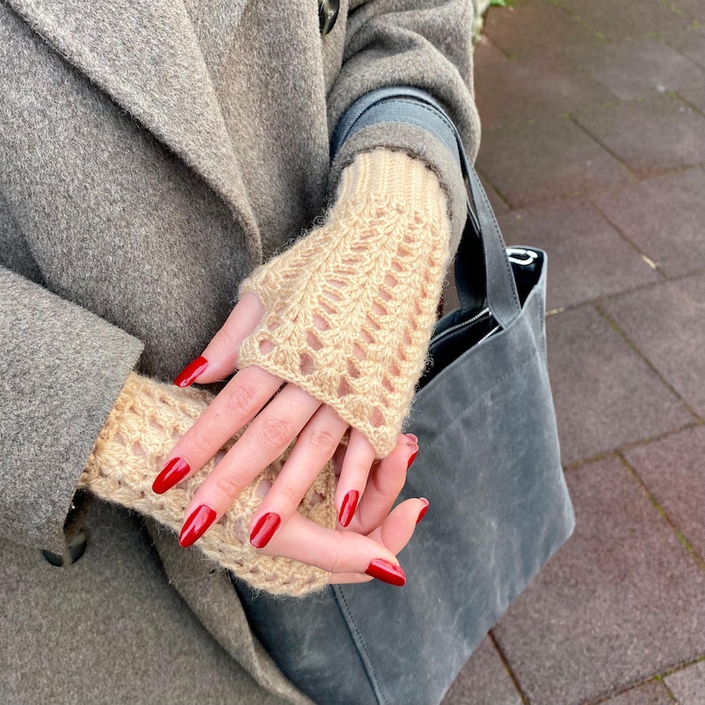 May include: A pair of beige fingerless gloves with a knitted openwork pattern. The gloves are worn with a grey coat and a grey tote bag. The person's nails are painted a bright red colour.