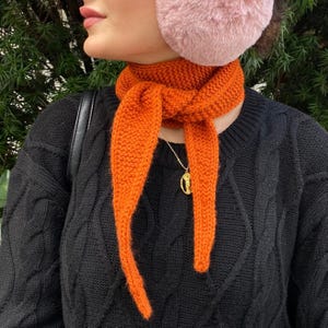 May include: An orange knitted scarf is tied around the neck, with long ends hanging down. The scarf is paired with a black cable-knit sweater and a gold pendant necklace. Pink earmuffs are worn on the head.