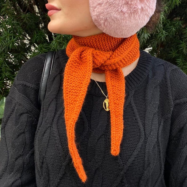 May include: An orange knitted scarf is tied around the neck, with long ends hanging down. The scarf is paired with a black cable-knit sweater and a gold pendant necklace. Pink earmuffs are worn on the head.