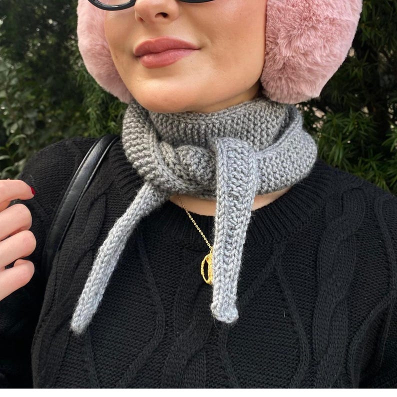May include: A gray knitted scarf tied around the neck, with long, dangling ends. The scarf is paired with a black sweater and pink earmuffs. A gold necklace is visible.