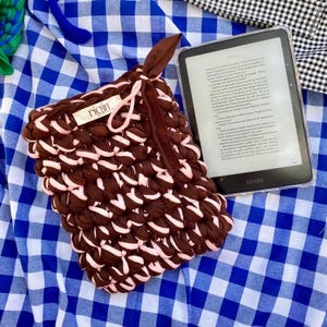 May include: A handmade brown and pink crocheted Kindle sleeve with a matching brown strap. The sleeve is made of thick, textured yarn and features a tag that reads "rigin". A Kindle e-reader is visible next to the sleeve.