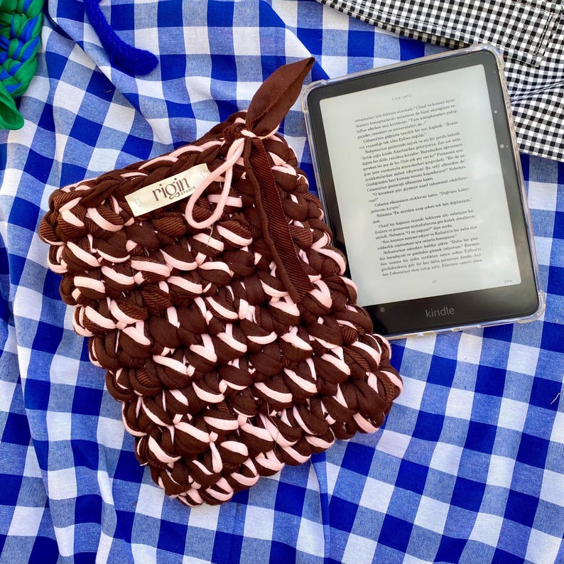 May include: A handmade brown and pink crocheted Kindle sleeve with a matching brown strap. The sleeve is made of thick, textured yarn and features a tag that reads "rigin". A Kindle e-reader is visible next to the sleeve.