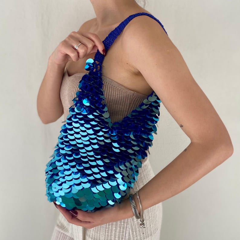 May include: A shoulder bag covered in overlapping, circular, metallic blue and teal sequins. The bag features a single, thick, crocheted blue strap that connects to the main body, which has a soft, slouchy shape and a shimmering, reflective surface.