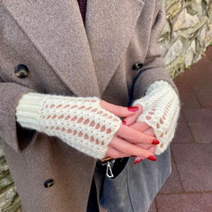 May include: Cream-coloured, open-weave fingerless gloves. The gloves are made of a soft, textured yarn and feature a delicate, lacy pattern. The wearer is also wearing a brown coat with black buttons.