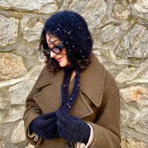 Könnte beinhalten: Ein dunkelblaues Strickmützen-, Schal- und Fäustlings-Set mit verstreuten Pailletten. Die Mütze hat ein Kapuzendesign, und der Schal hängt nach vorne. Die Fäustlinge sind in passendem Dunkelblau gehalten. Das Set wird mit einem braunen Mantel getragen.