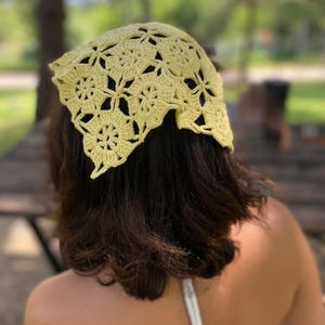 May include: A light yellow crochet headscarf with a floral pattern. The head covering is made of a soft, open-weave material. The headscarf is worn on the head.