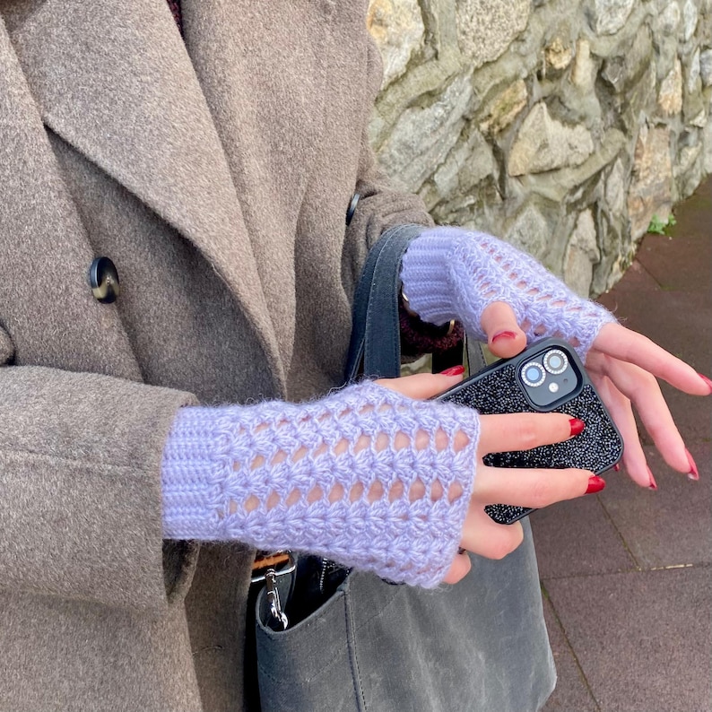 May include: Pale lilac fingerless gloves with an openwork pattern. The gloves are made of a soft, knitted material. They are worn with a grey coat and a black phone case.