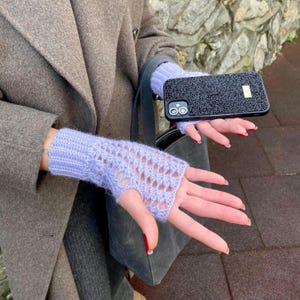 May include: Pale lilac fingerless gloves with a knitted openwork pattern. A black phone case with a textured finish and a gold-coloured detail is held in the hand. The gloves are made of a soft, knitted material.