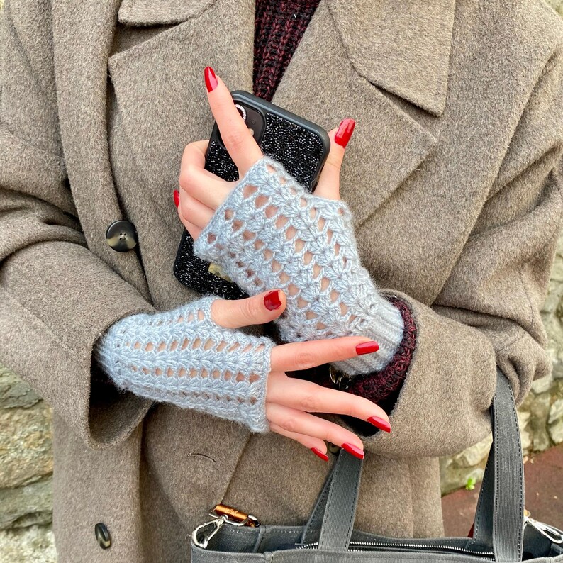 May include: Pale blue fingerless gloves with an openwork pattern. The gloves are knitted. The person is wearing a brown coat and holding a black phone. The coat has a button.
