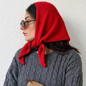 May include: A bright red knitted headscarf tied under the chin, paired with a dark grey cable-knit jumper and a tan handbag with brown leather straps. The headscarf features a ribbed texture and is worn over dark-rimmed sunglasses.