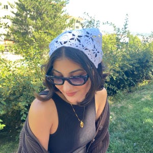 May include: A white crocheted headscarf with a floral pattern. The headscarf is worn on the head. The person is wearing sunglasses and a gold necklace. The background is a green, leafy outdoor setting.