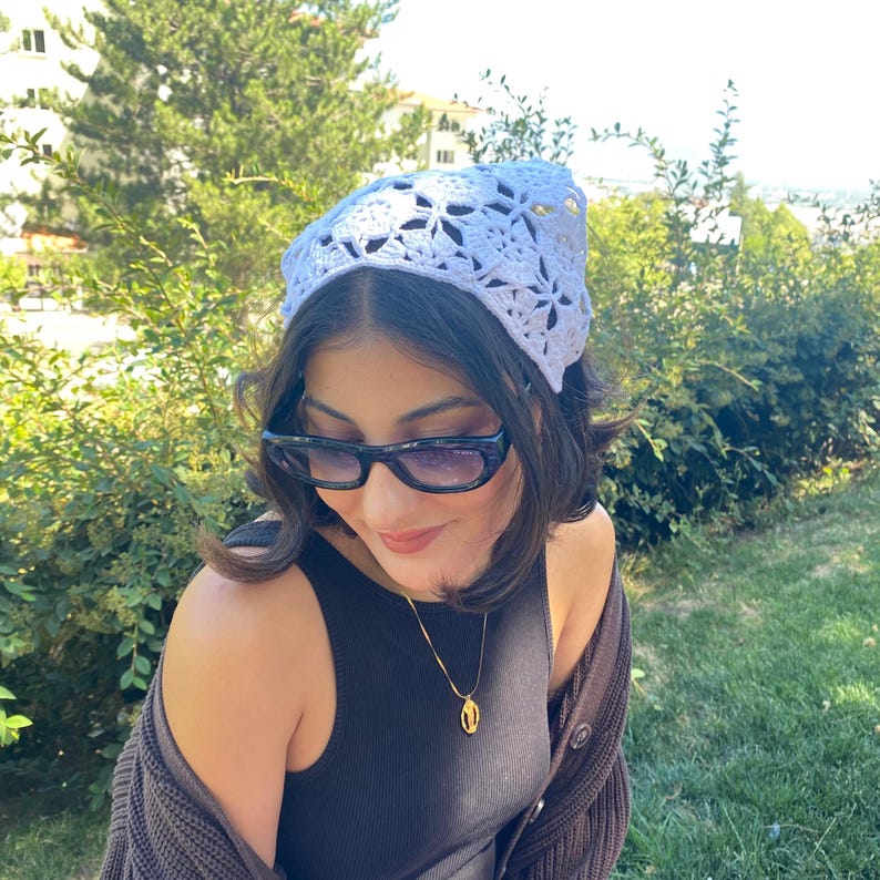 May include: A white crocheted headscarf with a floral pattern. The headscarf is worn on the head. The person is wearing sunglasses and a gold necklace. The background is a green, leafy outdoor setting.