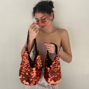 May include: Two hobo-style handbags crafted from black yarn and covered in large, reflective, metallic orange-red sequins. Each bag features a single integrated handle and a slouchy, rounded silhouette that showcases the shimmering scale-like texture.