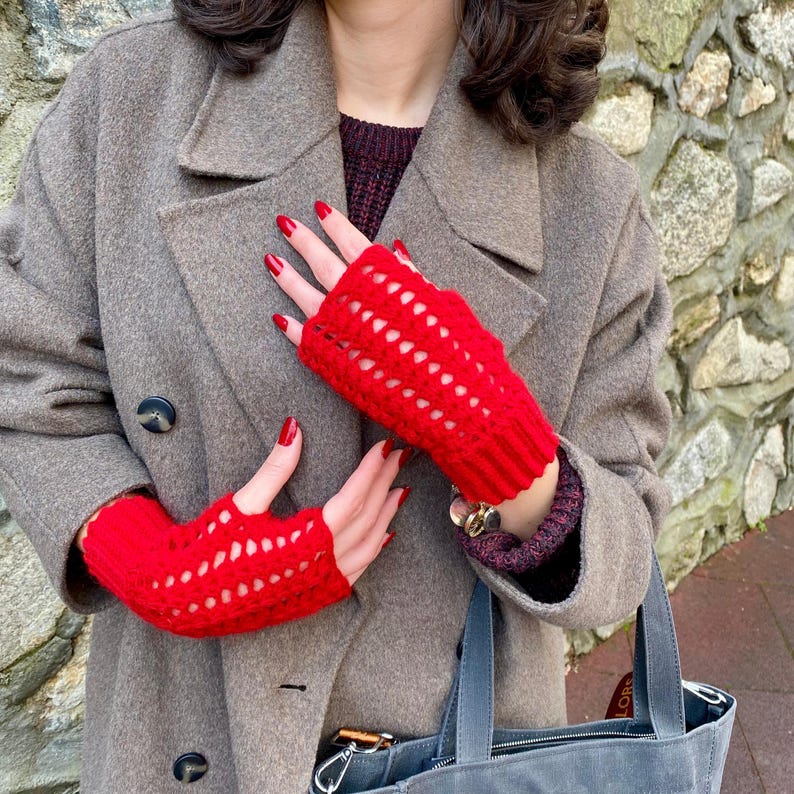 May include: Vibrant red fingerless gloves with a textured, open-weave pattern. The gloves are made of a soft, knitted material. The gloves are worn with a grey coat and a grey handbag.