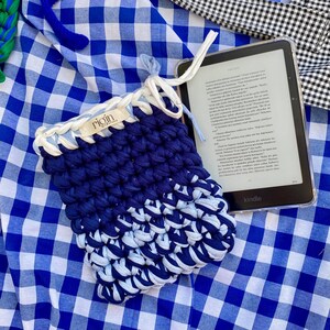 May include: A handmade blue and white crochet Kindle sleeve with the brand name "rigin" visible. The sleeve is made of thick, textured yarn and has a drawstring closure. The Kindle is visible inside the sleeve.