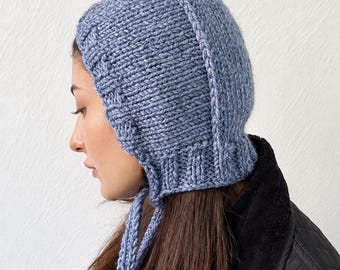 May include: A dusty blue knitted bonnet featuring a textured cable knit pattern along the front edge, a ribbed base, and two long knitted ties hanging down from the chin area. The bonnet is crafted from a thick, chunky yarn in a solid blue colour.