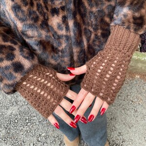May include: Brown fingerless gloves with a crocheted openwork design. The gloves are made of a soft yarn and are worn with a leopard print coat. The nails are painted red.