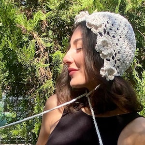 Peut inclure: Un chapeau en crochet crème avec des motifs floraux. Le chapeau présente un design ajouré délicat et est orné de plusieurs fleurs crochetées. Le chapeau a des liens blancs. Le fond est un feuillage vert luxuriant.