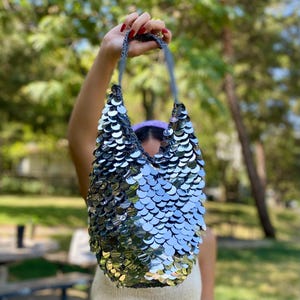 May include: A hobo-style shoulder bag covered in large, reflective silver sequins. The bag features a slouchy, unstructured silhouette with a single integrated strap, creating a shimmering, metallic appearance across the entire exterior surface.