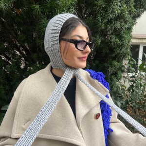 May include: A gray knitted hat with a long scarf attached. The hat is pulled down over the person's head and the scarf is tied around their neck. The person is wearing a beige coat and black sunglasses.