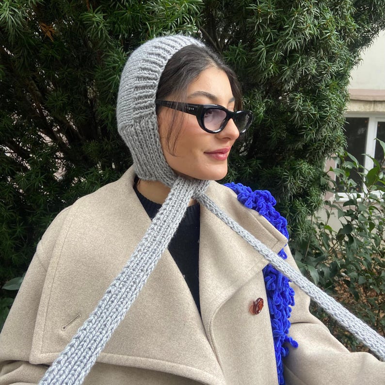 May include: A gray knitted hat with a long scarf attached. The hat is pulled down over the person's head and the scarf is tied around their neck. The person is wearing a beige coat and black sunglasses.