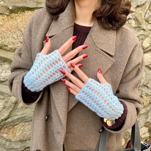 May include: Pale blue fingerless gloves with a delicate openwork pattern. The gloves are made of a soft, knitted material. The person is wearing a brown coat and a burgundy sweater.