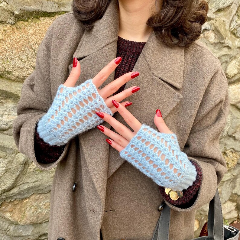 May include: Pale blue fingerless gloves with a delicate openwork pattern. The gloves are made of a soft, knitted material. The person is wearing a brown coat and a burgundy sweater.
