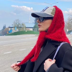 Può includere: Una persona indossa un cappuccio rosso sfocato e un berretto da baseball con una toppa. Il berretto ha una visiera grigia e una corona beige. La persona indossa anche occhiali da sole neri e un cappotto nero. Lo sfondo mostra una strada e un cielo nuvoloso.