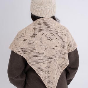 May include: A beige crochet triangular shawl featuring a large floral rose pattern in an openwork stitch design, paired with a matching beige knit beanie hat. The shawl has a scalloped edge and is worn over a dark brown coat.