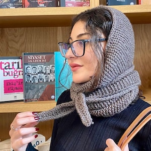 Könnte beinhalten: Graues Strick-Set aus Kapuze und Schal. Die Kapuze bedeckt Kopf und Hals, der Schal ist um den Hals gewickelt und gebunden. Das Set ist aus einem grob gestrickten Material. Im Hintergrund sind Bücher zu sehen.