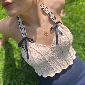 Può includere: Un top all'uncinetto color crema con bordo smerlato e dettagli in nastro nero. Il top presenta spalline decorative con dettagli intrecciati in bianco e nero e fiocchi neri. È visibile anche una collana con pendente dorato.