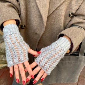May include: Light grey fingerless gloves with an open, lacy design. The gloves are knitted and have ribbed cuffs. They extend to the mid-forearm. The background shows a brown coat. The gloves are made of a soft material.
