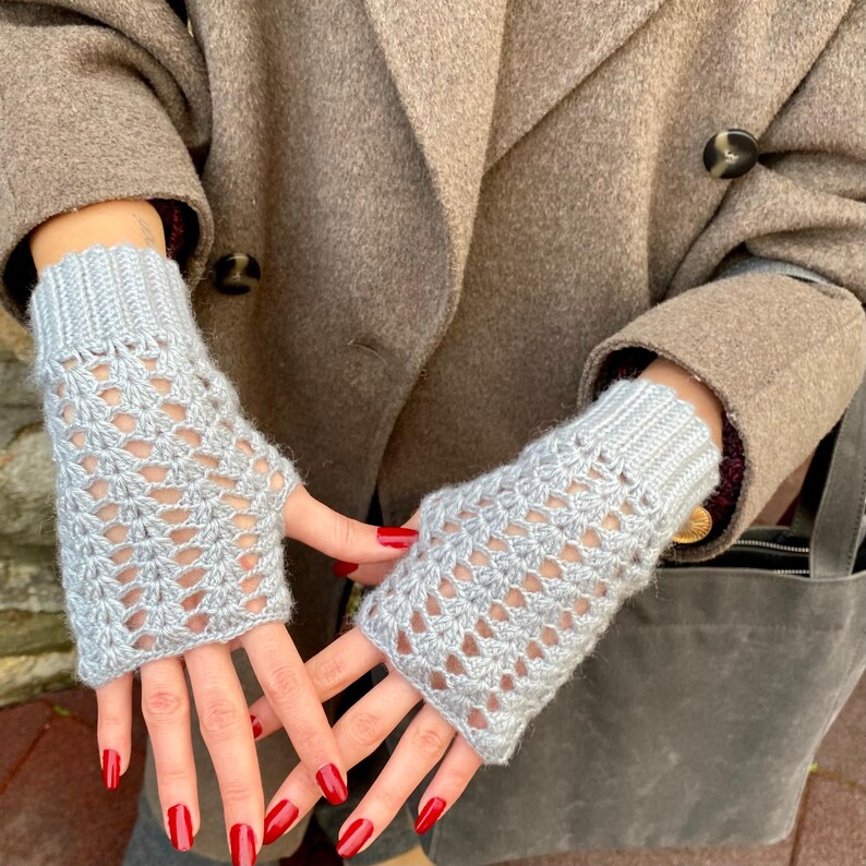 May include: Light grey fingerless gloves with an open, lacy design. The gloves are knitted and have ribbed cuffs. They extend to the mid-forearm. The background shows a brown coat. The gloves are made of a soft material.