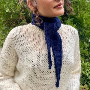 May include: A cream-colored, chunky knit sweater paired with a dark blue scarf. The scarf is knitted and hangs down the front. The sweater has a ribbed neckline and a loose, comfortable fit. The background is a blurred green foliage.