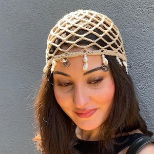 Könnte beinhalten: Ein beigefarbener Häkelkopfschmuck mit einem Rautenmuster. Der Kopfschmuck ist mit kleinen weißen Perlen, Muscheln und silberfarbenen Charms verziert. Der Kopfschmuck wird von einer Person mit dunklem Haar getragen.
