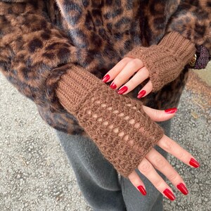 May include: Brown fingerless gloves with a knitted design. The gloves are made of a soft, textured material. The background includes a brown leopard print coat and grey trousers.