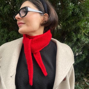 May include: A bright red knitted scarf is draped around a person's neck, contrasting with a black sweater and a light-colored coat. The scarf's ends hang down, creating a bold visual statement. The person is wearing black-framed glasses and a black headband.