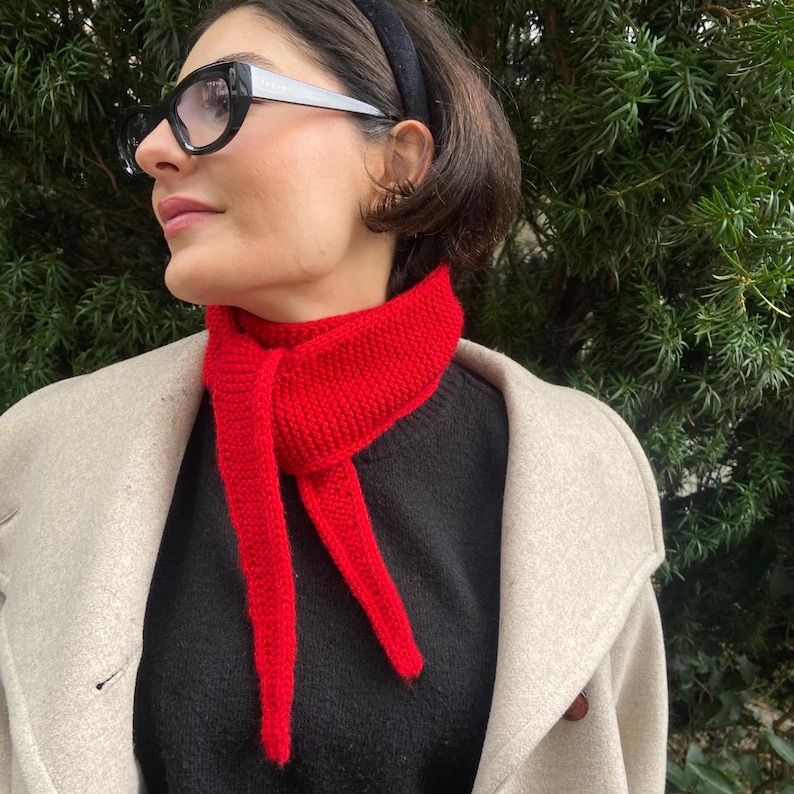 May include: A bright red knitted scarf is draped around a person's neck, contrasting with a black sweater and a light-colored coat. The scarf's ends hang down, creating a bold visual statement. The person is wearing black-framed glasses and a black headband.