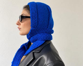 May include: A vibrant royal blue knitted balaclava hood with a thick, textured garter stitch pattern. The headpiece features a structured hood design that extends into a scarf-like wrap around the neck, crafted from chunky yarn in a solid blue colour.