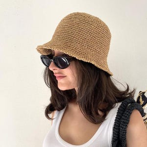 Könnte beinhalten: Ein beigefarbener Häkel-Fischerhut mit texturiertem Webmuster und einer kurzen, leicht nach unten geneigten Krempe. Der Hut wird mit einer schwarzen, ovalen Sonnenbrille und einem weißen Tanktop getragen, dazu ist ein schwarzer Taschenriemen sichtbar.
