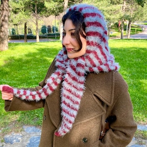 Puede incluir: Bufanda con capucha a rayas rojas y grises. La bufanda est&aacute; hecha de un material suave y difuso y se usa sobre un abrigo marr&oacute;n. El dise&ntilde;o presenta rayas rojas y grises alternas, creando un accesorio visualmente llamativo.