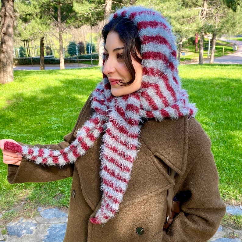 Puede incluir: Bufanda con capucha a rayas rojas y grises. La bufanda est&aacute; hecha de un material suave y difuso y se usa sobre un abrigo marr&oacute;n. El dise&ntilde;o presenta rayas rojas y grises alternas, creando un accesorio visualmente llamativo.