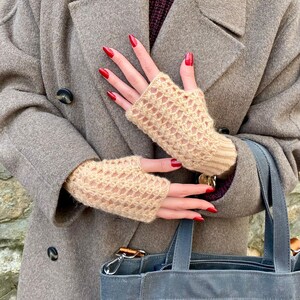 May include: Beige fingerless gloves with a knitted openwork pattern. The gloves are worn with a grey coat and a blue tote bag. The person's nails are painted red.