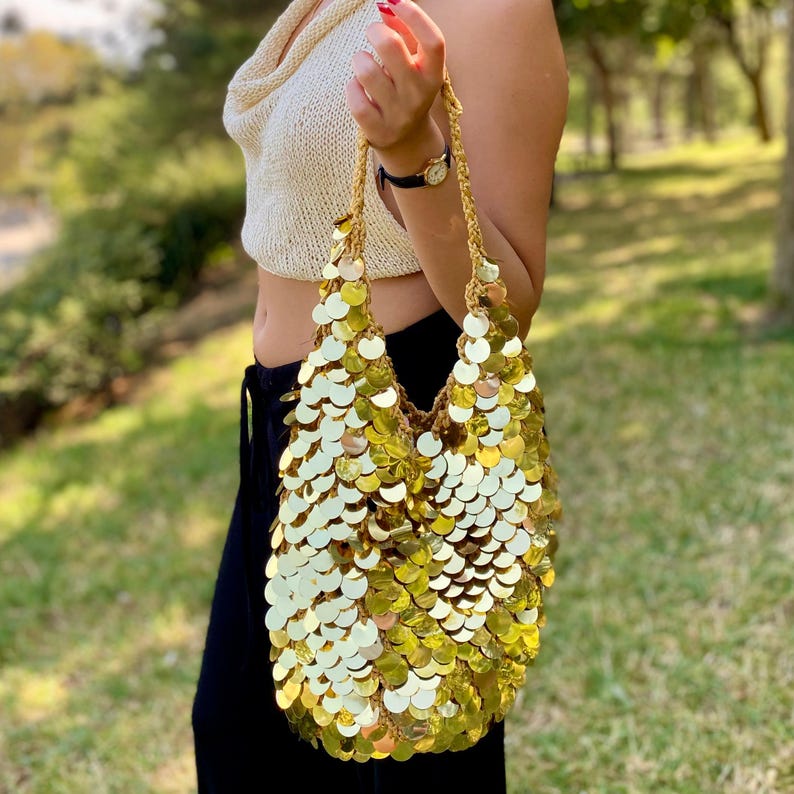 May include: A hobo-style shoulder bag covered in large, reflective gold sequins. The bag features a crocheted base structure with a braided strap, creating a shimmering, scale-like texture across the entire exterior surface of the accessory.