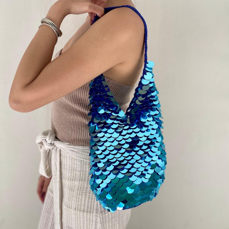 May include: A hobo-style shoulder bag covered in overlapping, circular, metallic blue and teal sequins. The bag features a single crocheted strap in a matching deep blue yarn, creating a textured, scale-like appearance across the entire surface.