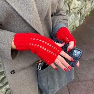 May include: Vibrant red fingerless gloves with a crocheted pattern. The gloves are made of a soft, textured material. The person is wearing a grey coat and holding a black phone case.