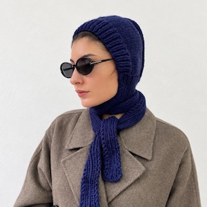 Könnte beinhalten: Eine dunkelblaue Stricksturmhaube mit integriertem Schal, der um den Hals gewickelt ist. Die Kopfbedeckung hat eine gerippte Textur und wird mit einer runden schwarzen Sonnenbrille und einem beigen zweireihigen Wollmantel getragen.