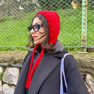 May include: A bright red knit hood with long ties, worn with a black coat and sunglasses. The hood is a vibrant, solid color, and the ties hang down the front. The overall look is stylish and warm, suitable for cooler weather.