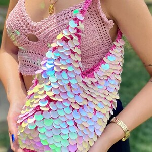 May include: A crochet shoulder bag crafted from vibrant pink yarn, completely covered in iridescent, circular sequins that shimmer with a pearlescent finish. The bag features a single wide strap and a structured, slouchy silhouette with a textured base.