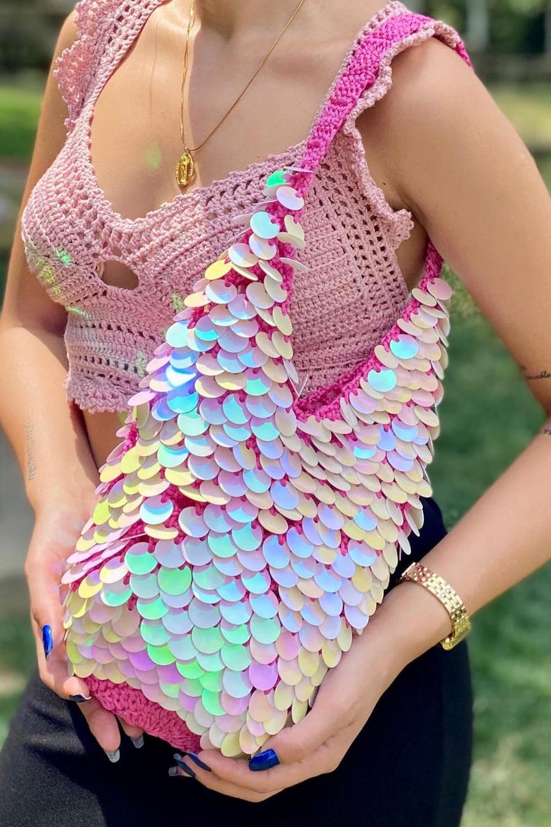 May include: A crochet shoulder bag crafted from vibrant pink yarn, completely covered in iridescent, circular sequins that shimmer with a pearlescent finish. The bag features a single wide strap and a structured, slouchy silhouette with a textured base.